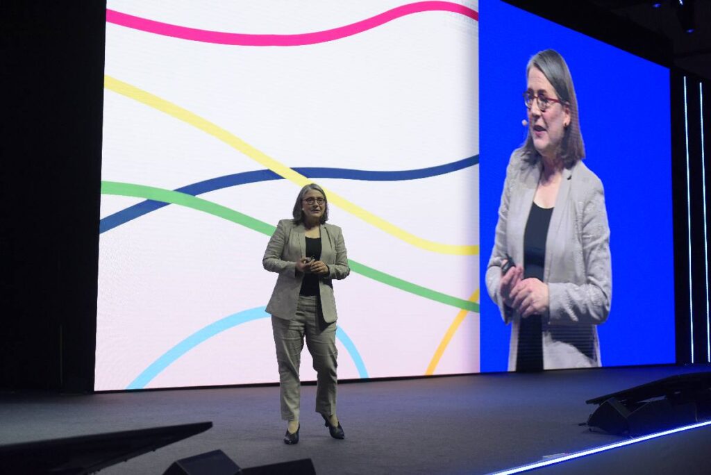 Woman with glasses (Michele Wucker) speaking on a stage against a white backdrop with colored wavy lines and giant-sized video of the woman.