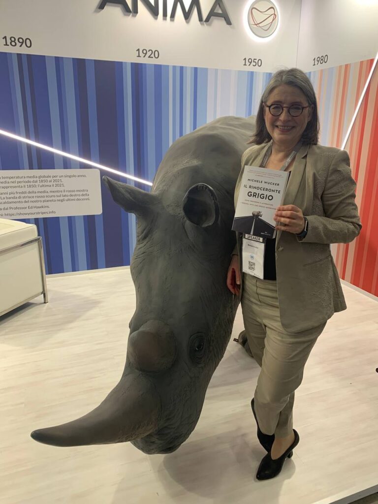 A woman holding a copy of the book "Il Rinoceronte Grigio" next to a large resin rhinoceros against a backdrop of a graphic showing rising earth temperatures from 1890 to 1980; and the name and logo for ANIMA Asset Management.